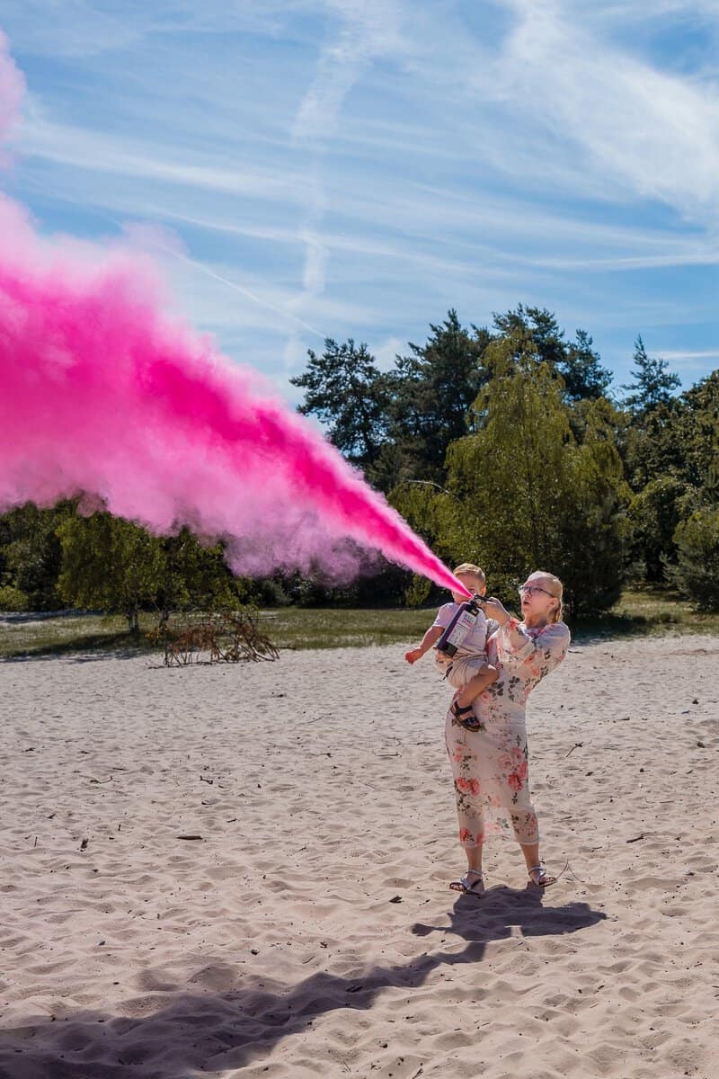 Moeder met zoontje spuit een wolk roze poeder met een gender reveal brandblusser in de duinen
