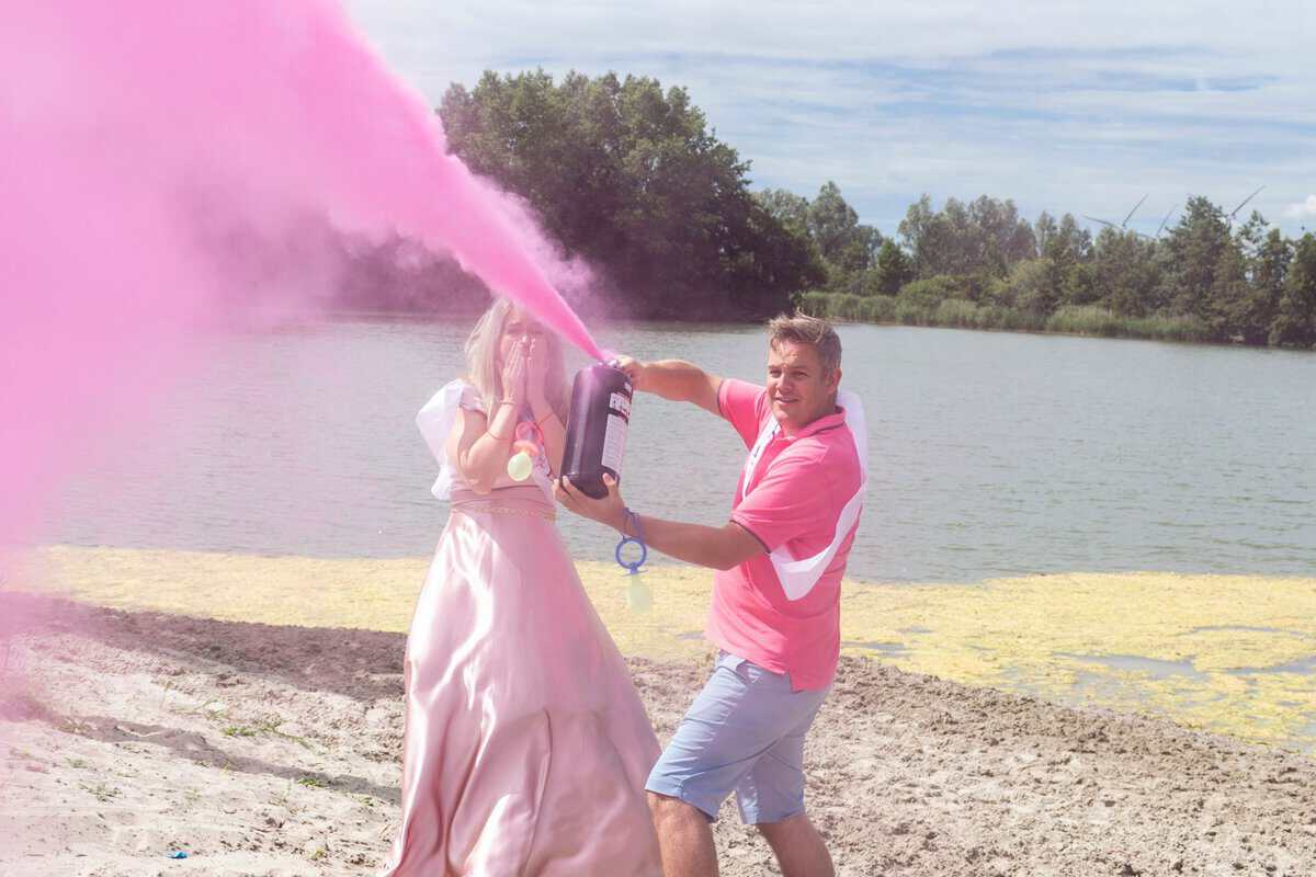 Man spuit een grote roze wolk uit een gender reveal brandblusser bij een meer, vrouw met handen voor haar mond